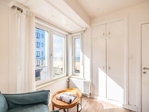 Een kamer met uitzicht op zee, een raam met zonwering, een radiator, een houten tafel met een tijdschrift en een groene bank.