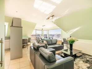 A modern living room with a sofa set, a glass coffee table, and a view of the cityscape through large windows. - Bright penthouse with roof terrace near the sea (Koksijde)