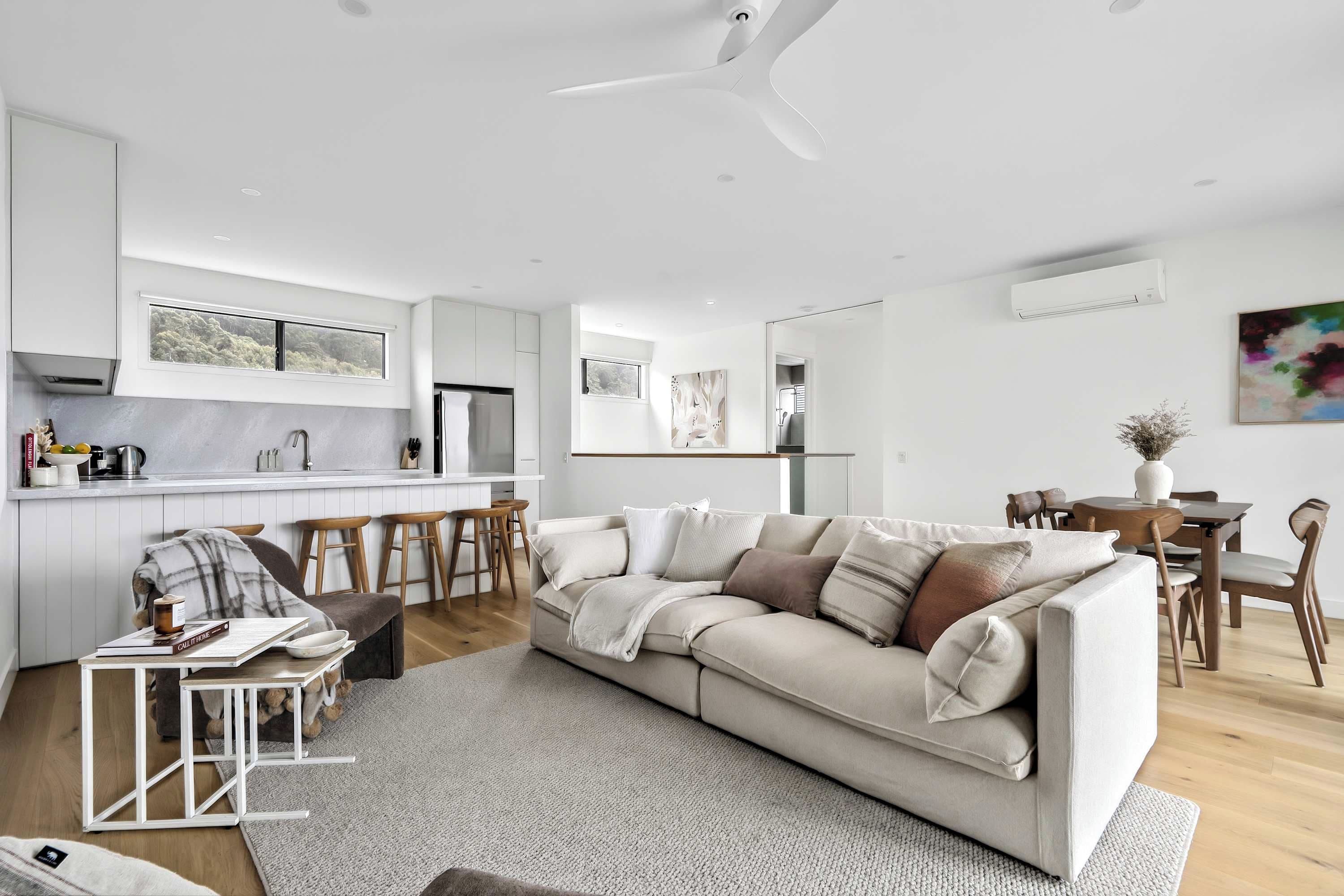 Una sala de estar moderna con un sofá color beige, una zona de comedor con sillas de madera y una cocina con armarios blancos.