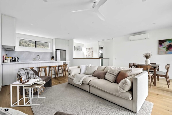 Een moderne woonkamer met een beige bank, een eethoek met houten stoelen en een keuken met witte kastjes.