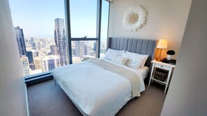 A bedroom with a large bed, a nightstand with a lamp, and a view of a cityscape through a large window. - Luxury Panoramic Views - 2B2B Pool, Gym & Parking (Melbourne)