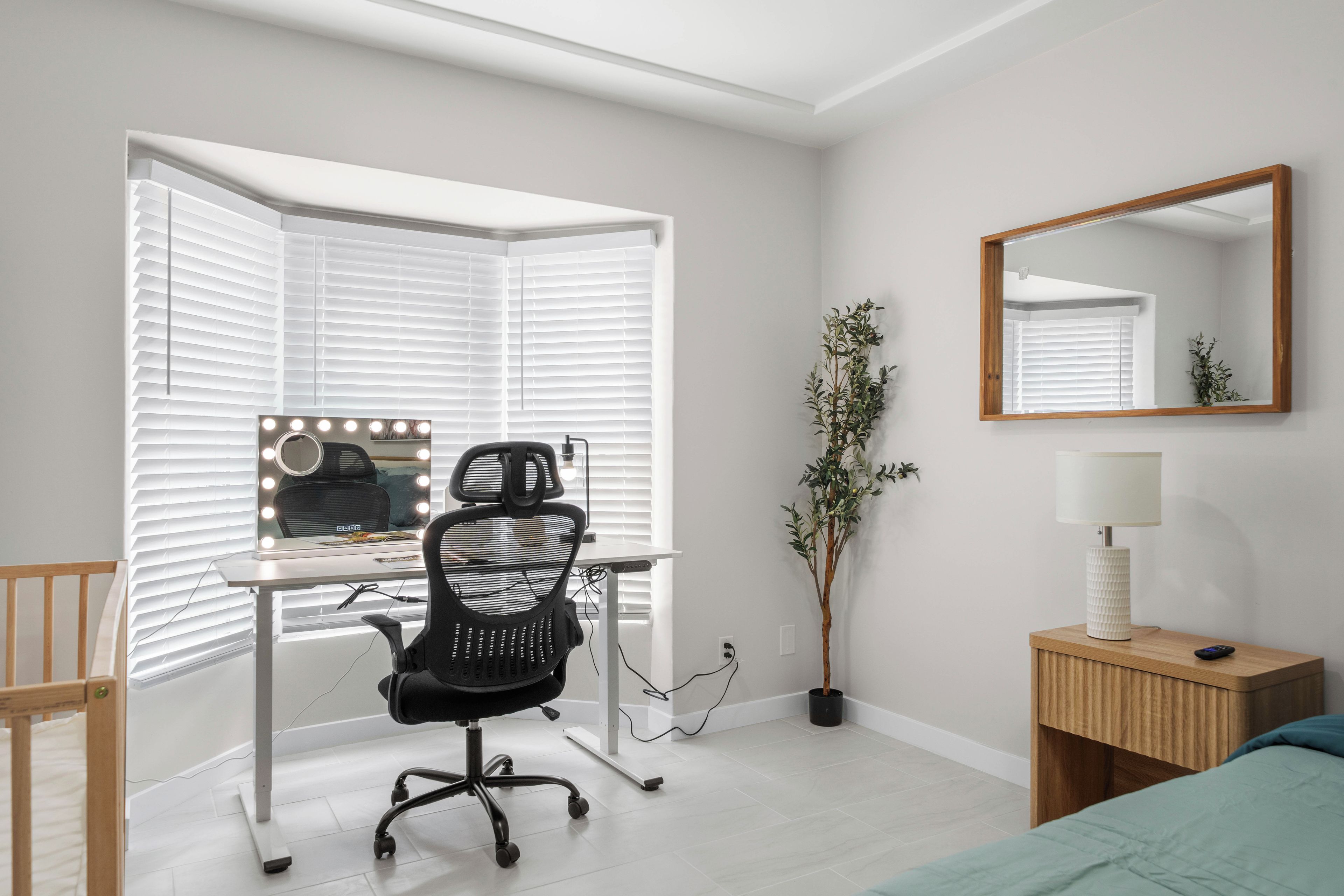 A bedroom with a desk, chair, mirror, bedside table, lamp, and a potted plant.