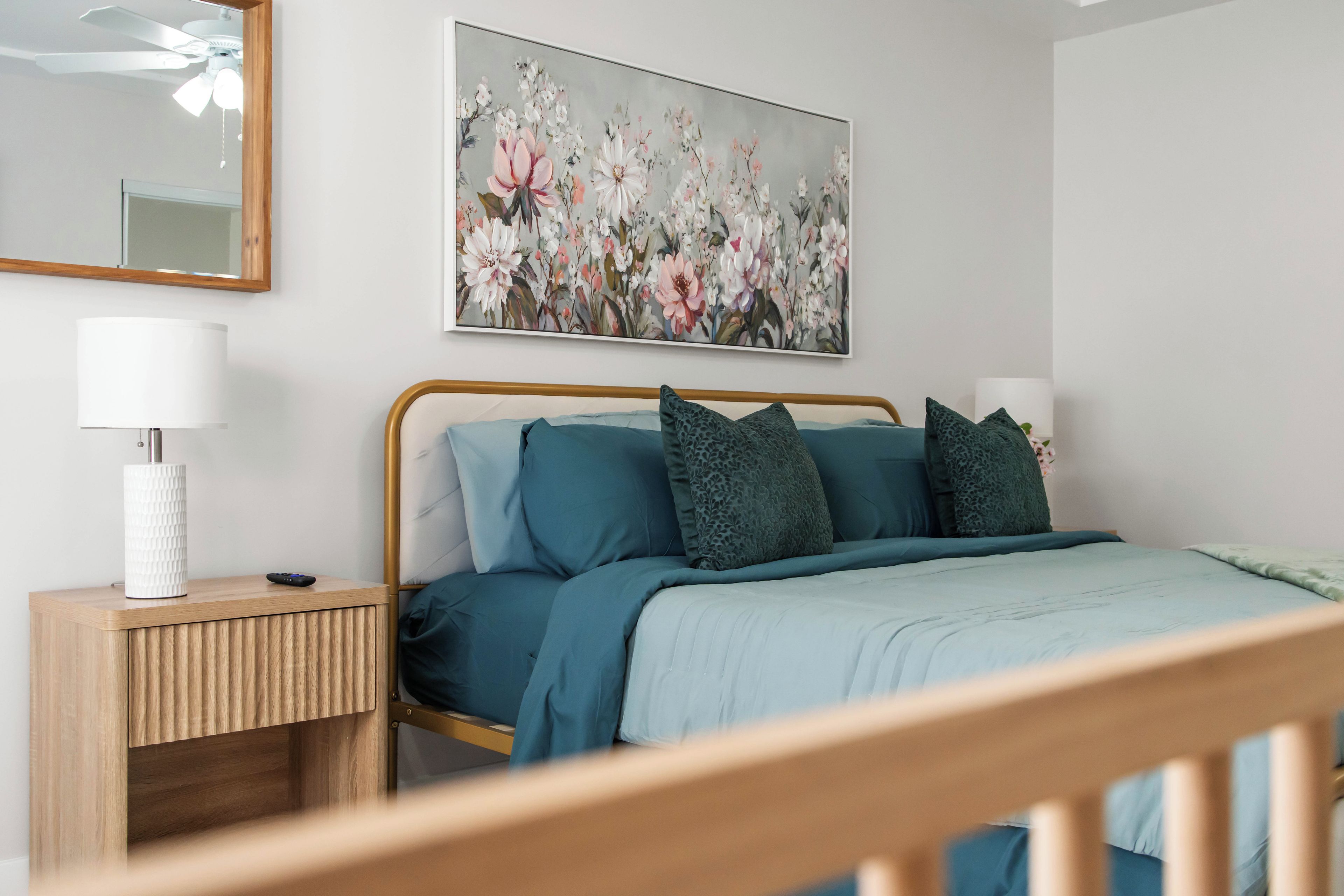 A bedroom with a bed, a bedside table, a lamp, a mirror, and a floral painting on the wall.