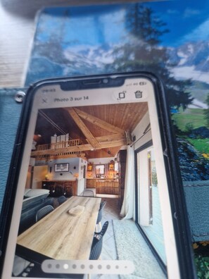 A smartphone displaying a photo of a room with a wooden dining table, chairs, and a kitchen area with wooden cabinets and a hanging light fixture. - New Listing (Aubessagne)