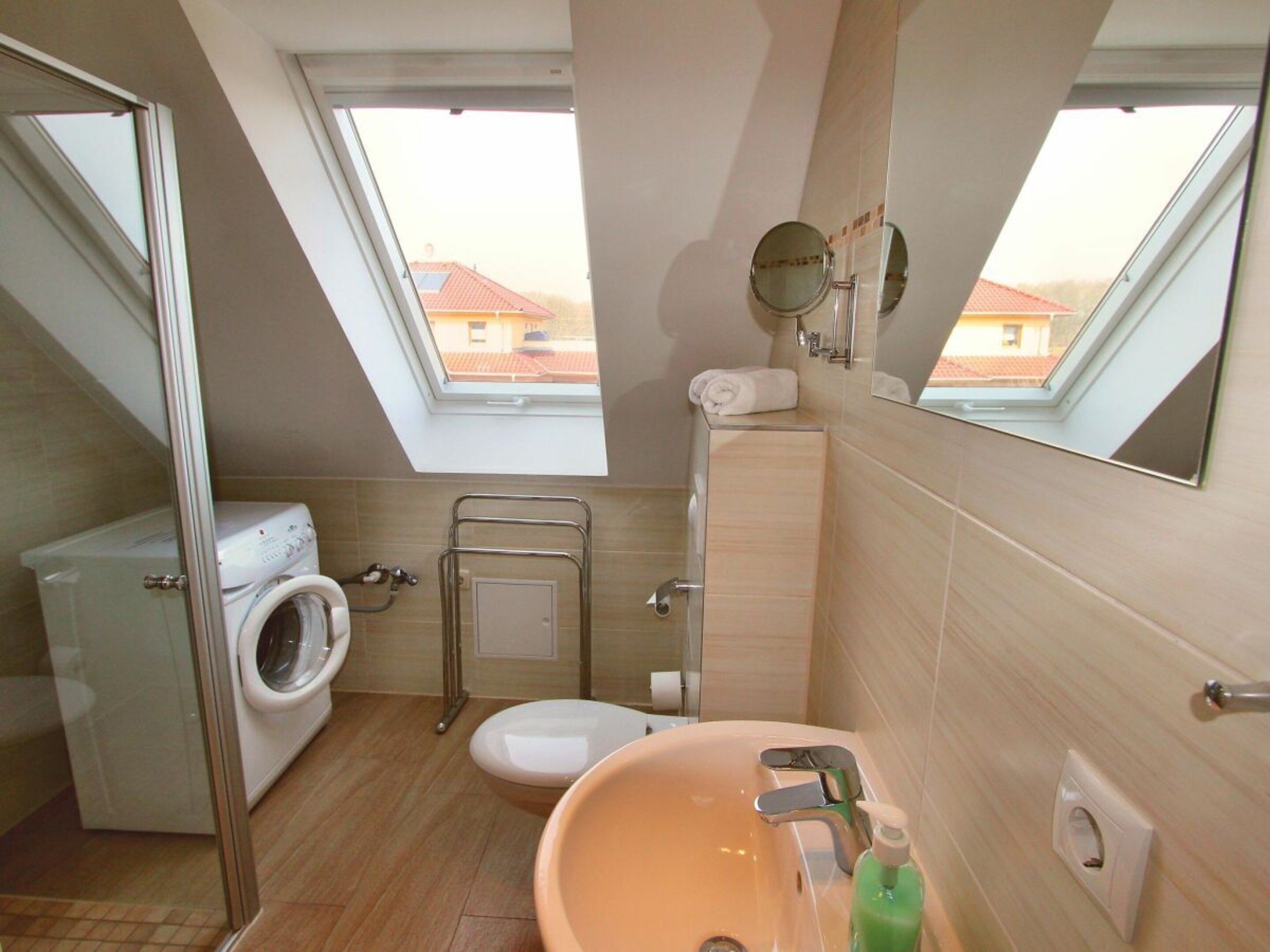 A compact bathroom with a washing machine, toilet, and sink, featuring skylights and a view of buildings outside.