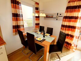 Un comedor con una mesa dispuesta para cuatro, sillas, una ventana con cortinas y una zona de cocina al fondo.