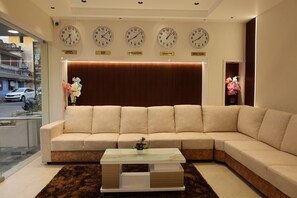 A modern living room with a beige sectional sofa, a glass coffee table, and wall clocks displaying time from various cities. - The Golden Park (Dindigul)