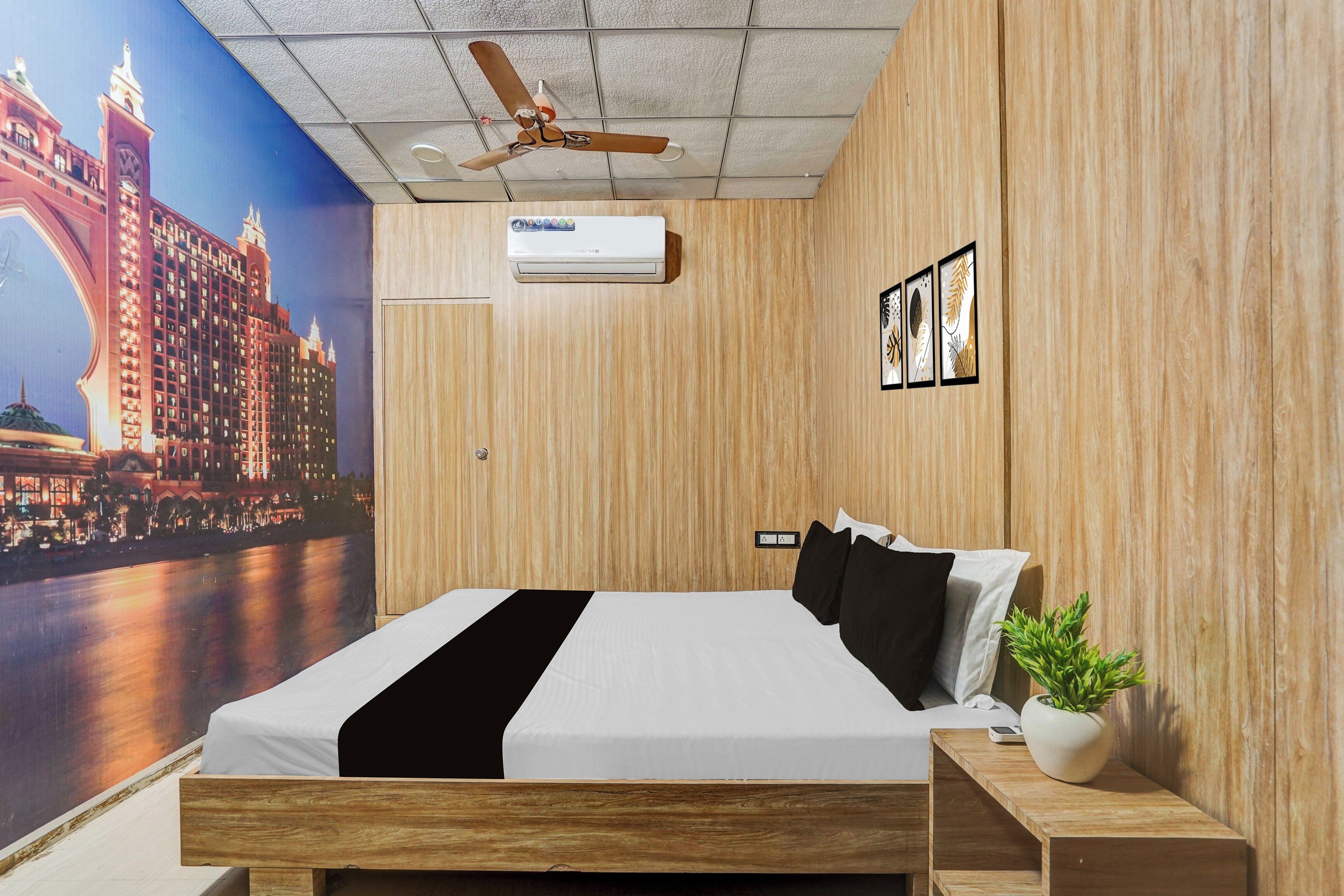 A hotel room with a bed, a ceiling fan, a wall-mounted air conditioner, a nightstand with a plant, and a mural of a cityscape.