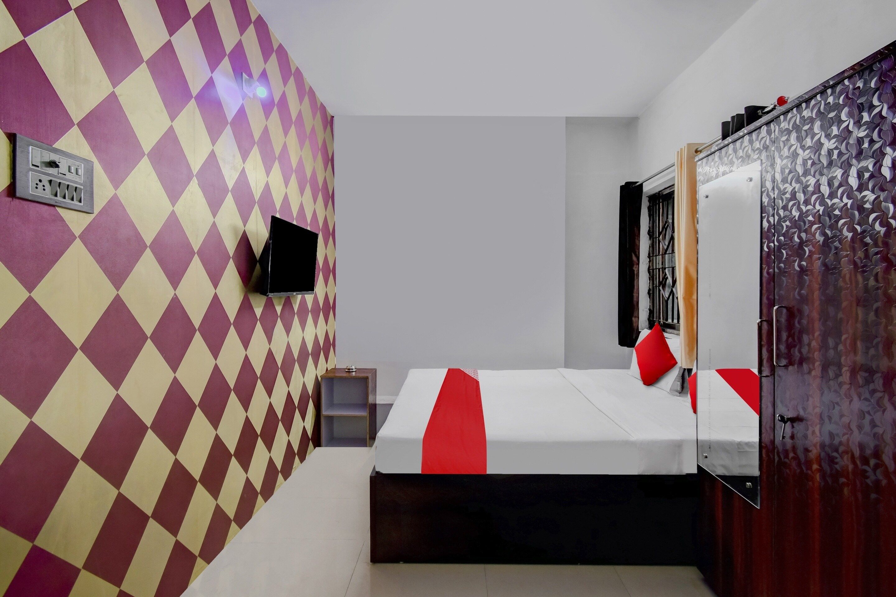 Quarto de hotel com padrão quadriculado na parede, cama com detalhes vermelhos, mesa de cabeceira pequena, TV fixada na parede e guarda-roupa com porta estampada.