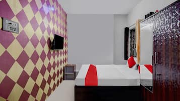 Quarto de hotel com padrão quadriculado na parede, cama com detalhes vermelhos, mesa de cabeceira pequena, TV fixada na parede e guarda-roupa com porta estampada.