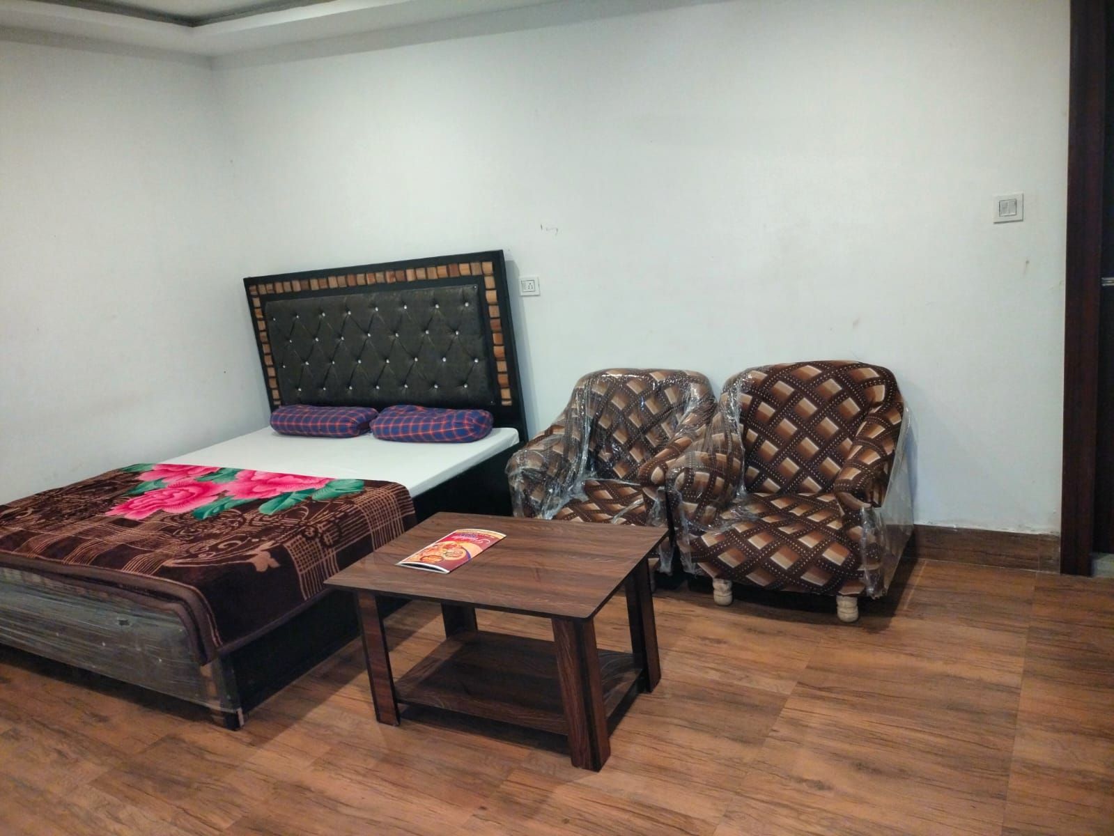Quarto com cama, duas poltronas, uma mesinha de madeira e parede branca.