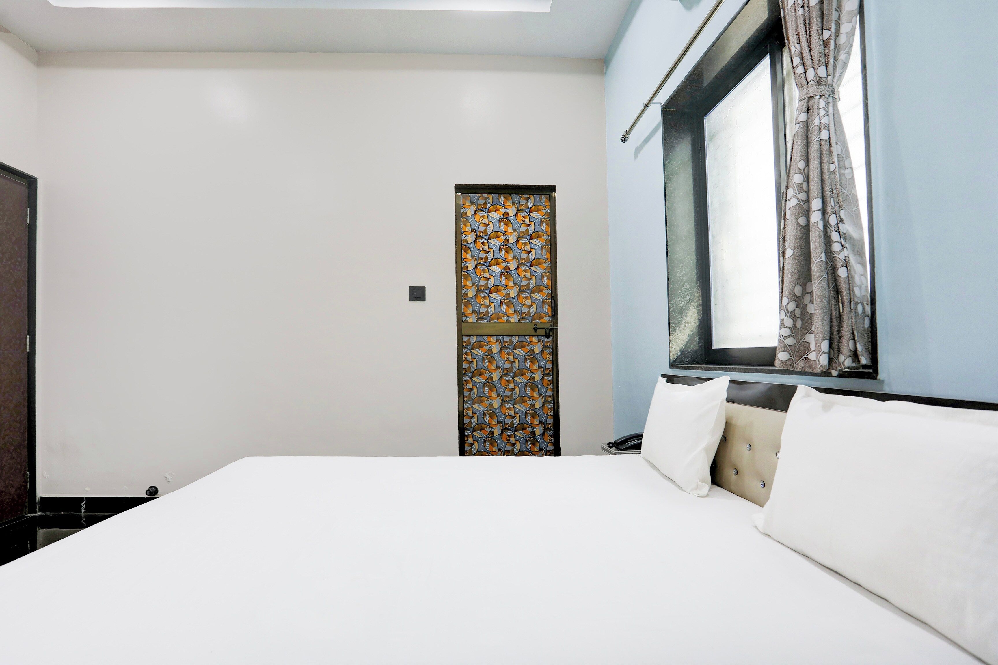 Une chambre d’hôtel avec un lit, une fenêtre avec des rideaux, une porte ornée de motifs et un interrupteur sur le mur.
