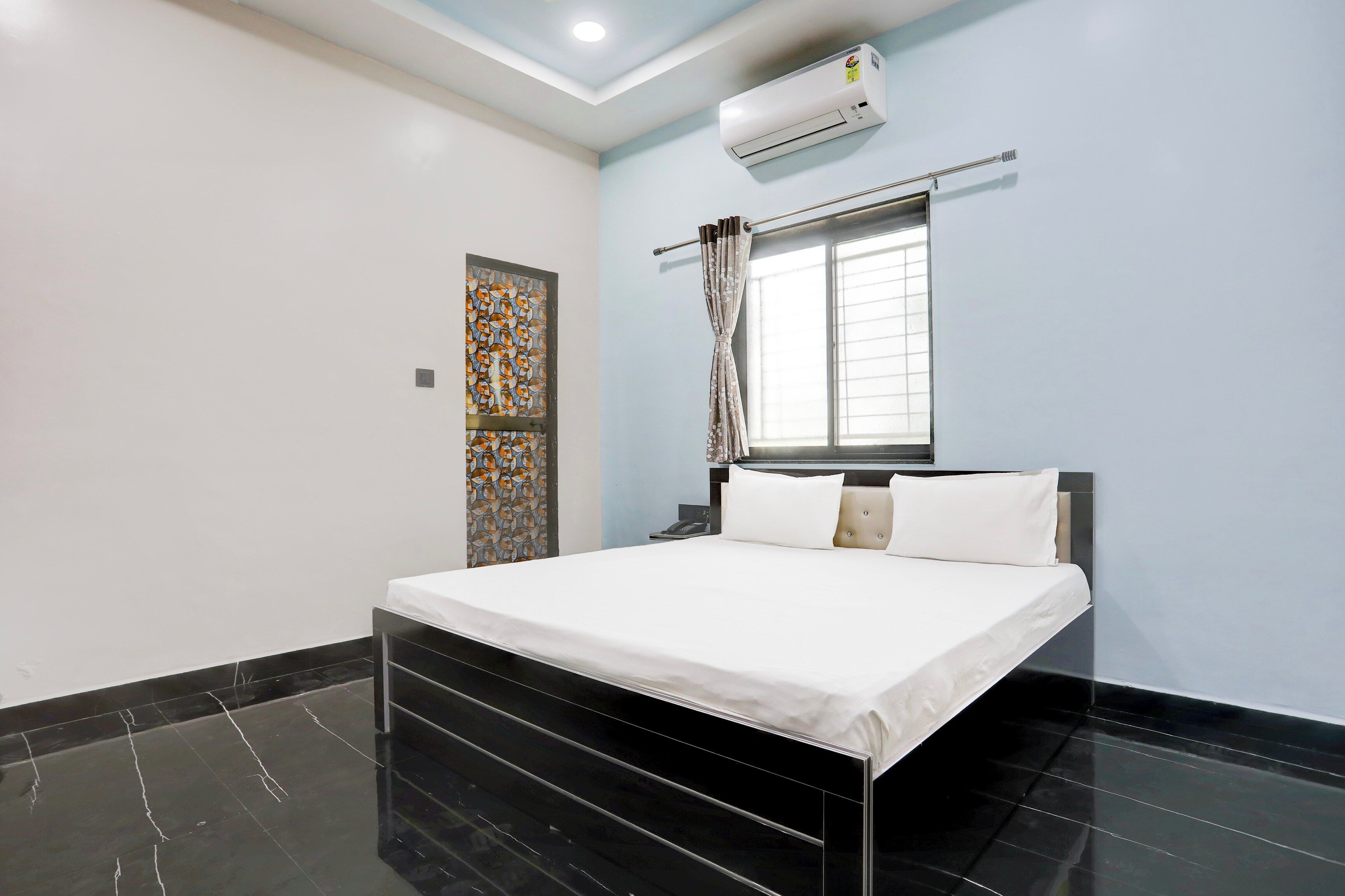 Una habitación de hotel con cama, ventana con cortinas, equipo de aire acondicionado y puerta decorativa.