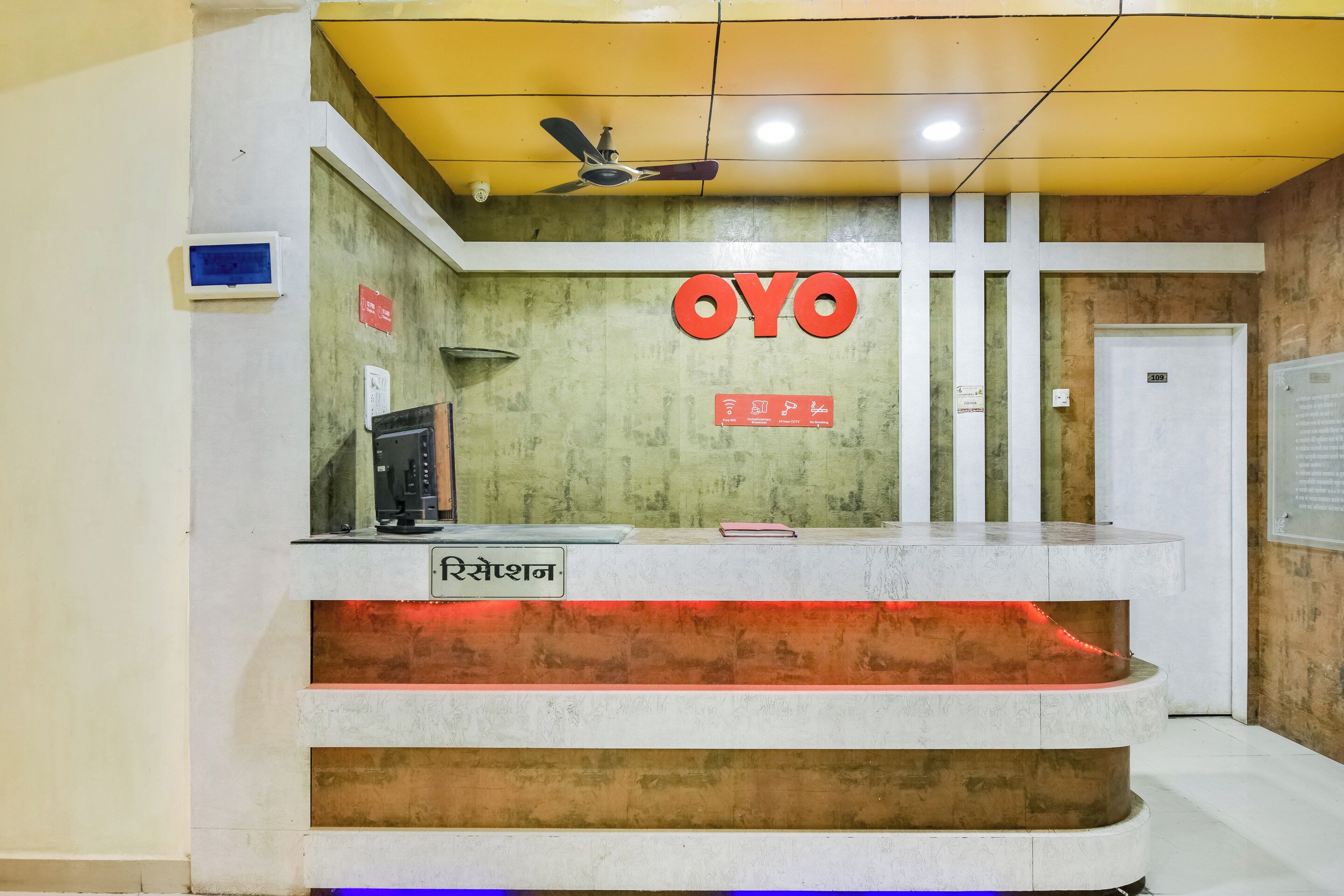 Un mostrador de recepción con un ventilador de techo, un letrero con la marca 'OYO' y una pantalla digital.