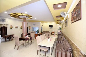 A dining area with multiple tables and chairs, a large pillar, ceiling fans, and a television mounted on the wall. - Hotel O H.K.Continental (Amritsar)