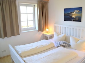 Ein Schlafzimmer mit Bett, Nachttisch, Lampe, einem Fenster mit Vorhängen und einem Bild an der Wand.