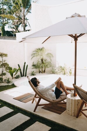 A person relaxing on a wicker lounge chair under a white umbrella in a well-maintained outdoor patio area with a pool, surrounded by various plants and a white wall. - ASHITA by SEAOM Villas (Gianyar)