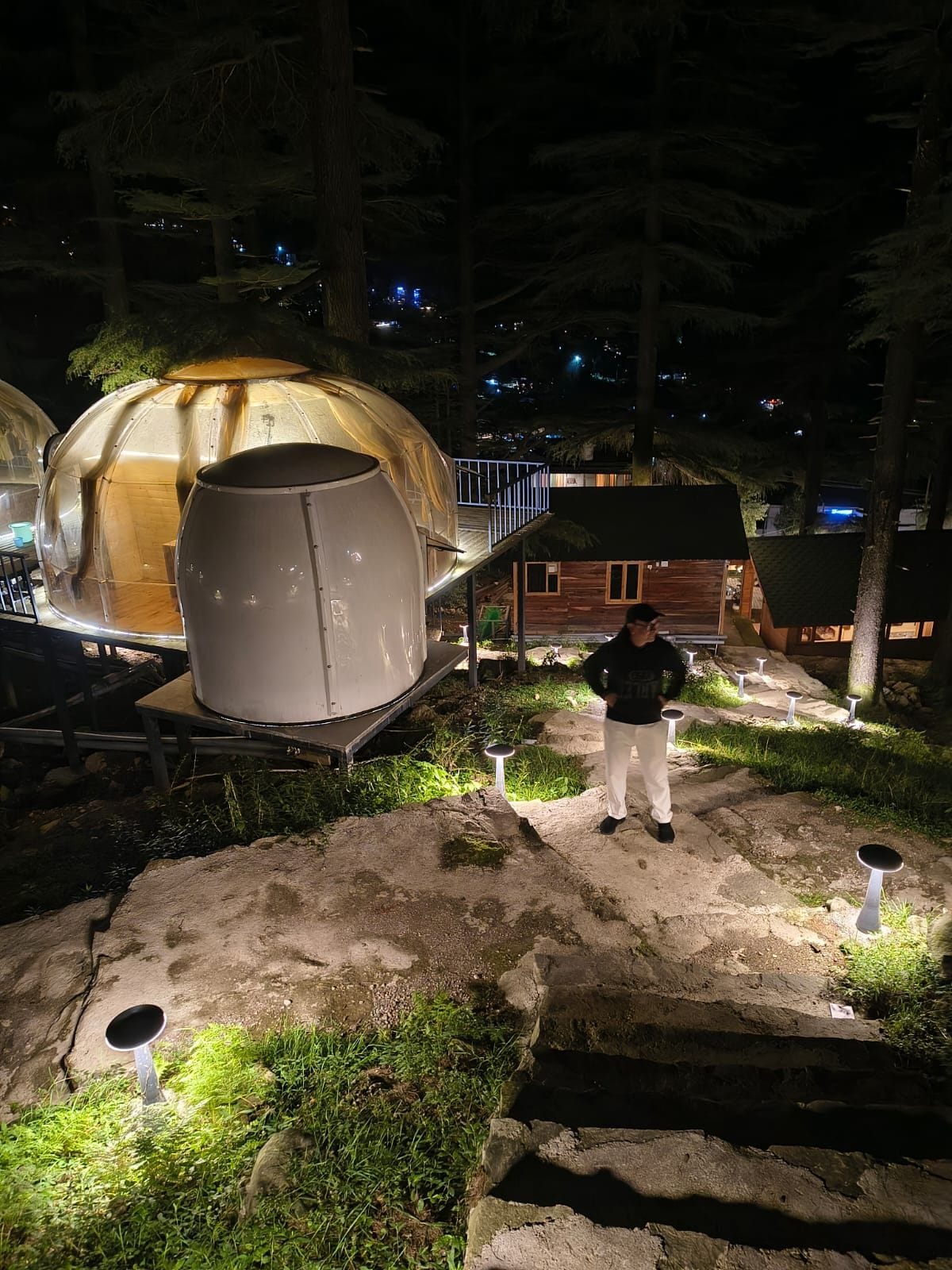 A dome-shaped structure with a cylindrical object beside it, a wooden cabin, and a person standing in the foreground.