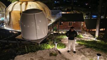 A dome-shaped structure with a cylindrical object beside it, a wooden cabin, and a person standing in the foreground.