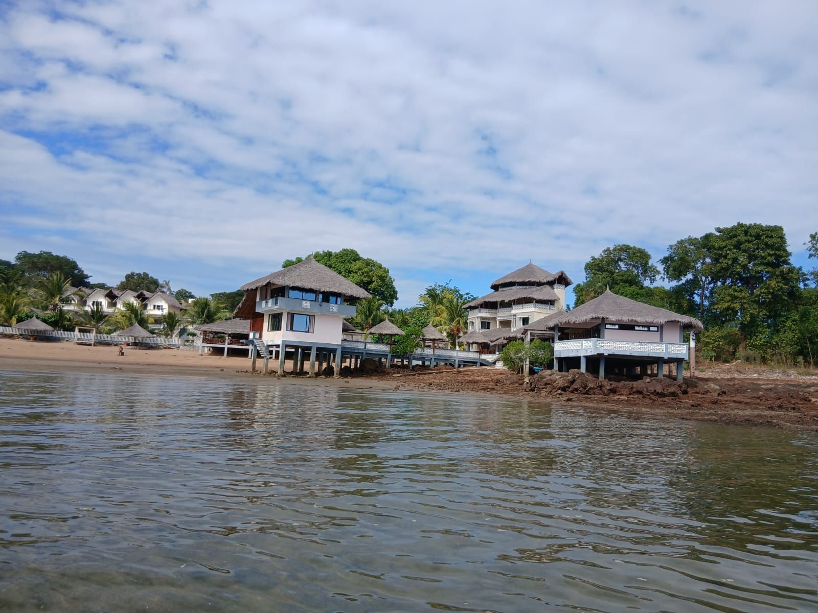Une propriété située en bord de mer, comprenant plusieurs bâtiments, dont certains sont dotés de toits de chaume, et une plage de sable en premier plan.
