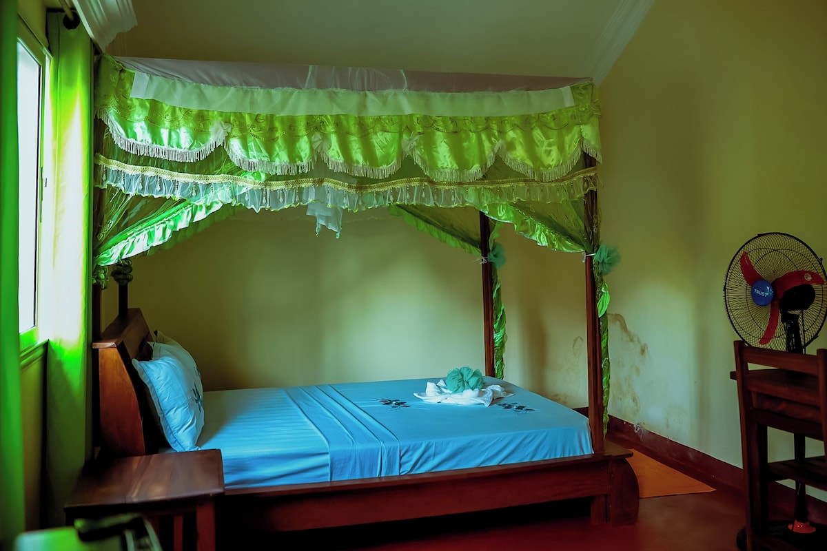 Una habitación con cama, un banco de madera, un ventilador de pared y una ventana con cortinas.