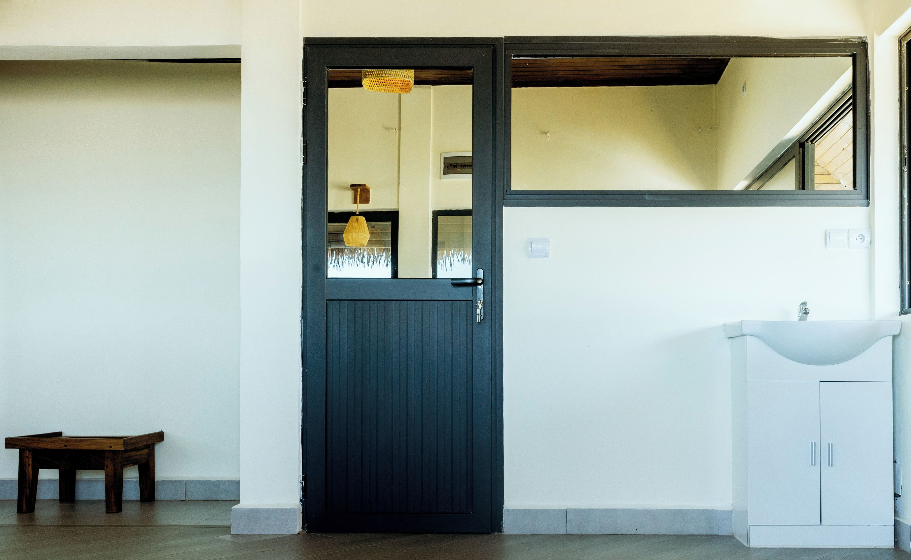 Une pièce avec une porte sombre, un petit banc en bois, un lavabo blanc et un miroir au mur.