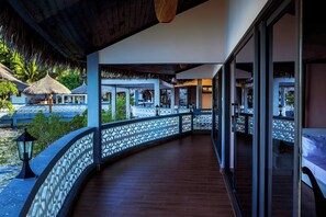 Un espacio al aire libre techado con piso de madera curvo, barandas decorativas blancas y vistas a un resort tropical al fondo.