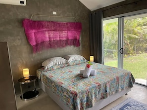A bedroom with a bed, pillows, a blanket, bedside lamps, and a view of greenery outside.