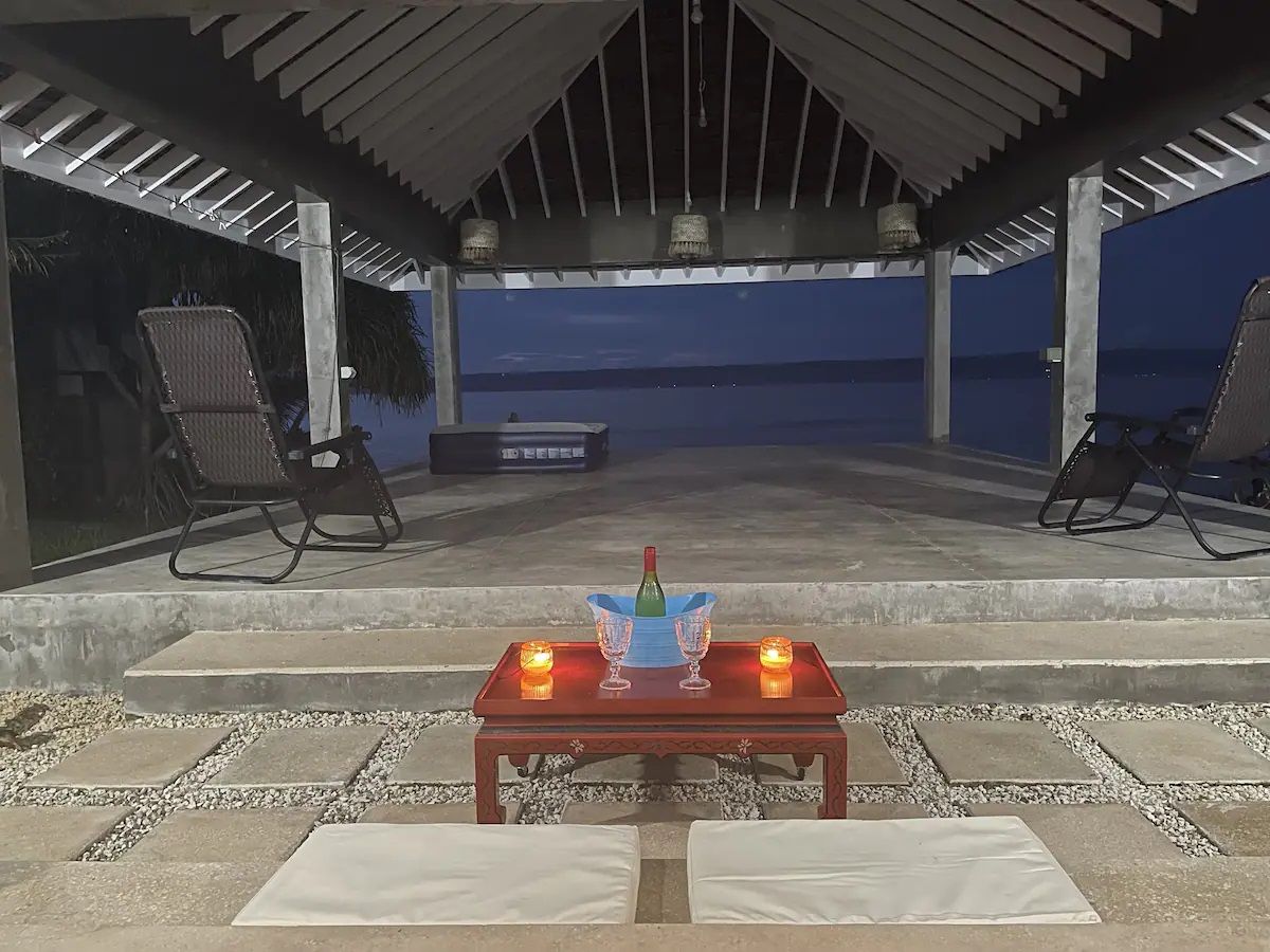 An outdoor covered patio with a table set for two, featuring a bottle and glasses, and two chairs facing a view.