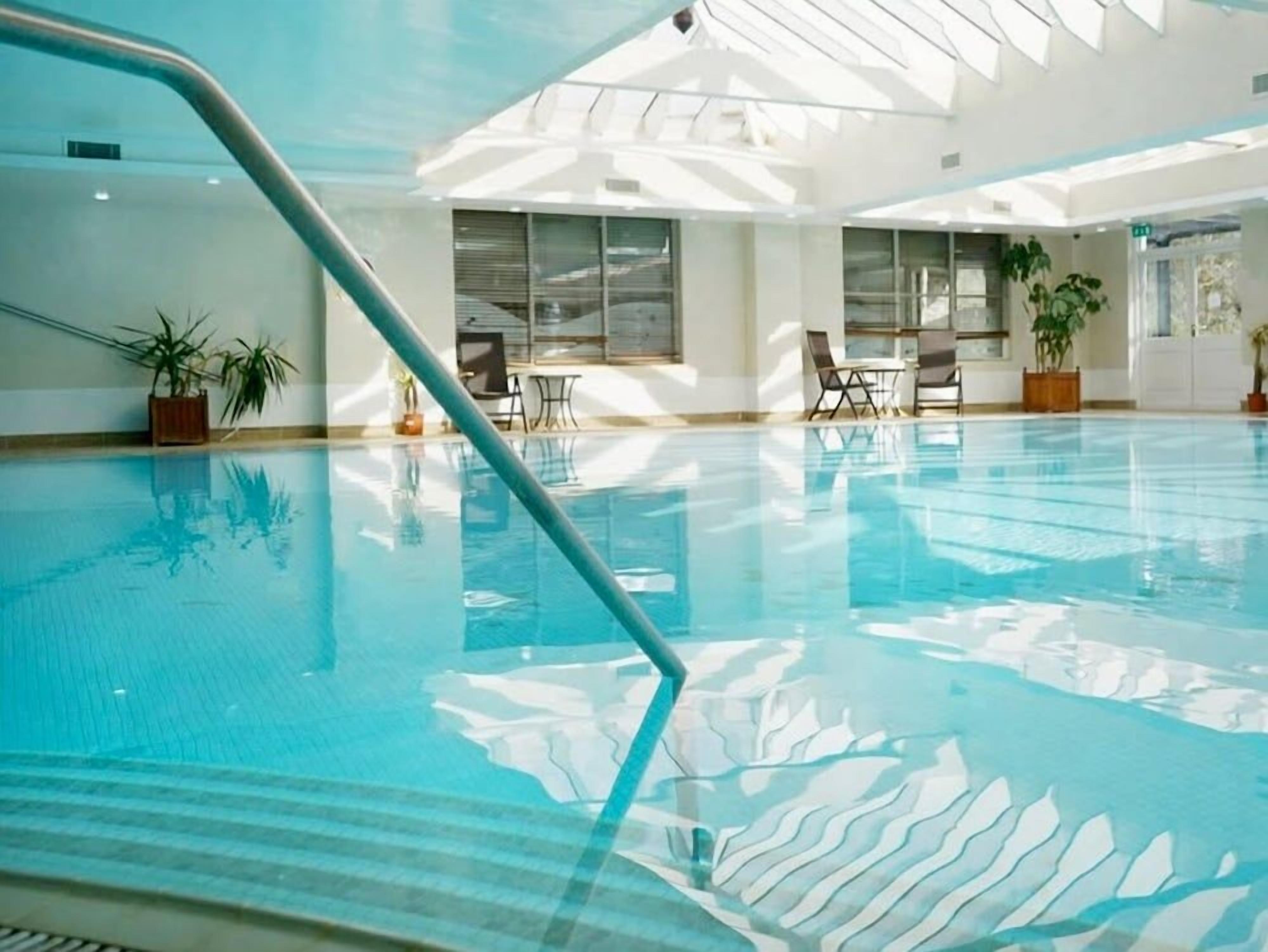 Una piscina interior con agua cristalina, ambiente iluminado por el sol y sillones de descanso.