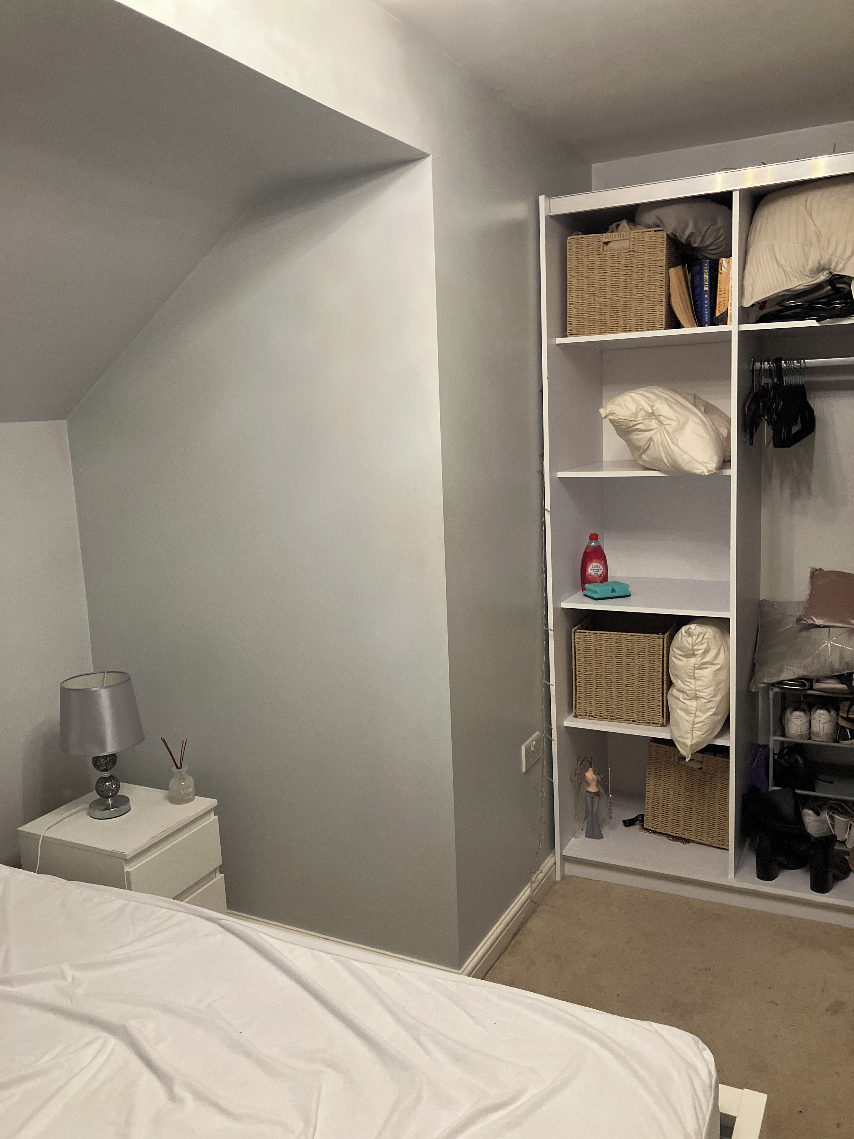 A bedroom with a bed, a nightstand with a lamp, a wardrobe with shelves, and a carpeted floor.