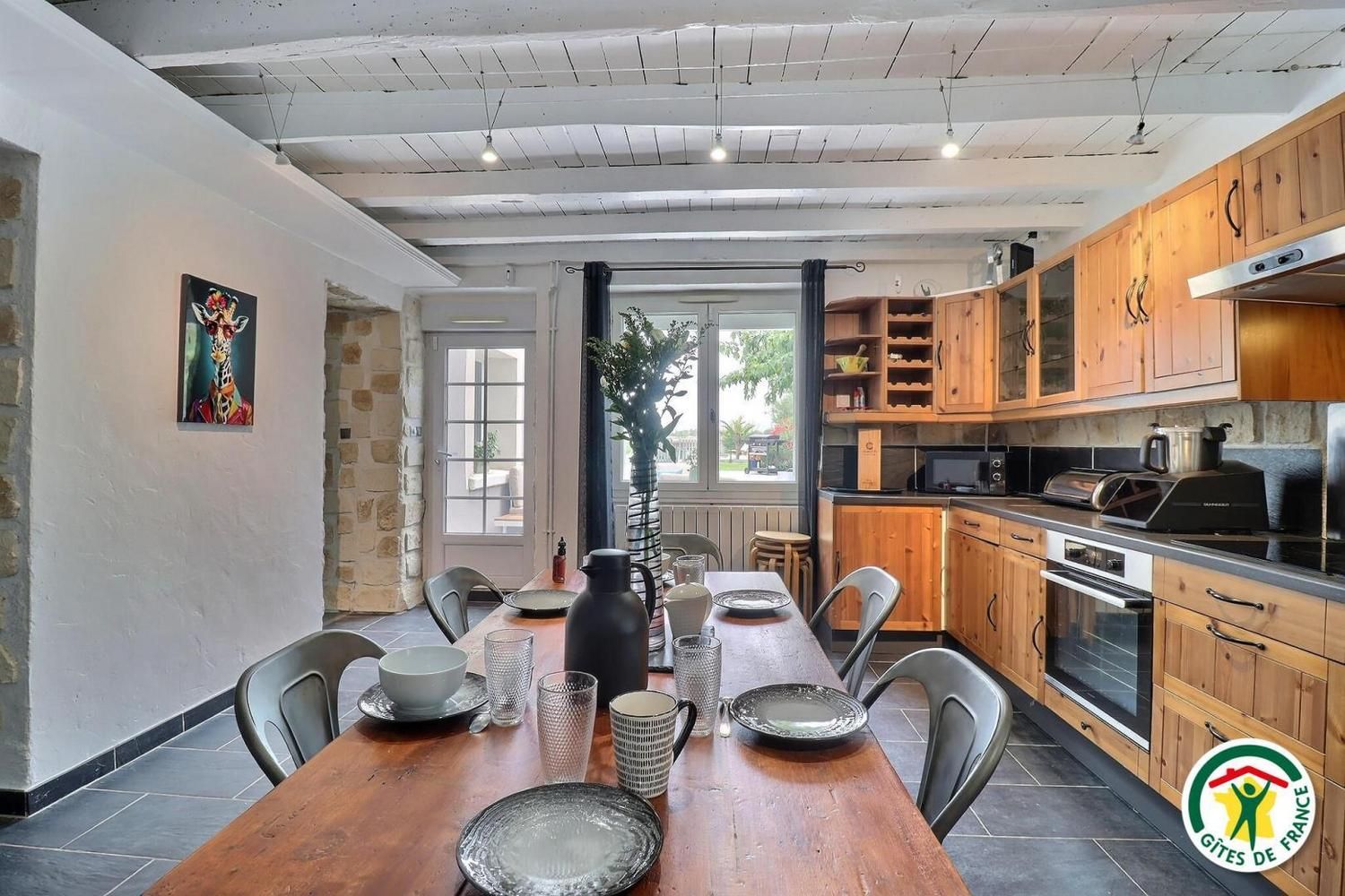 Une cuisine moderne avec des meubles en bois, une table à manger dressée avec des assiettes, des verres et des couverts, et une vue sur l’extérieur grâce à une grande fenêtre.