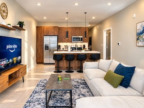 A modern living room with a flat-screen TV, a sofa, a coffee table, and a kitchen area with bar stools. - Magic City Oasis | 2 King 1 Queen 2B (Birmingham)