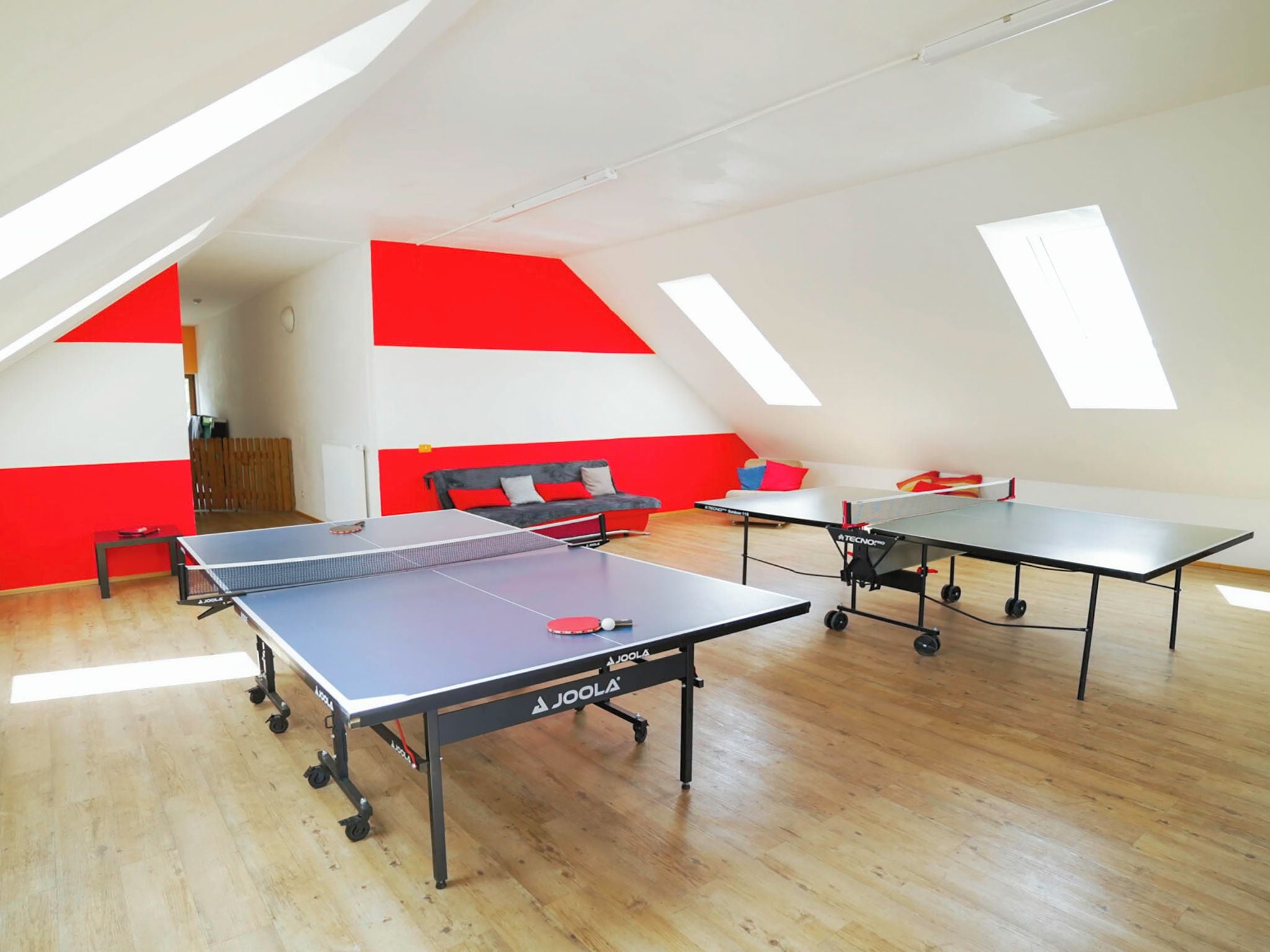 Amplia habitación con dos mesas de ping pong, un sofá y una cama, con techo inclinado y tragaluces.