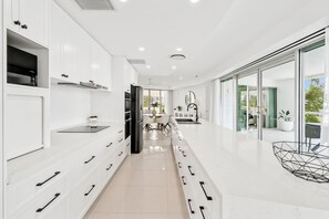 A modern kitchen with white cabinetry, a central island, and a dining area with a view. - River Esplanade Penthouse (Mooloolaba)