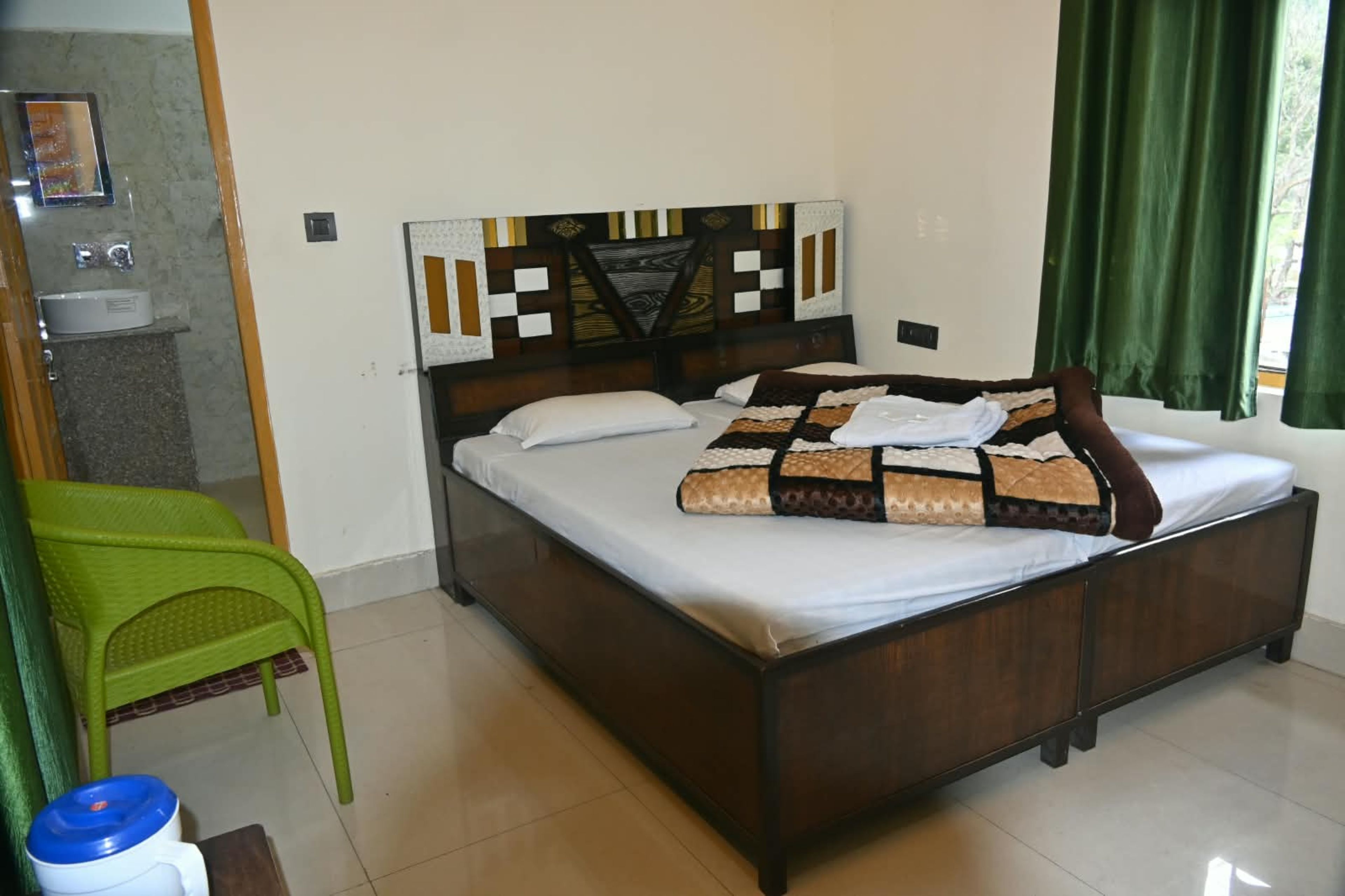 Una cama individual con colcha estampada, una mesita de noche, una silla verde y una ventana con cortinas.