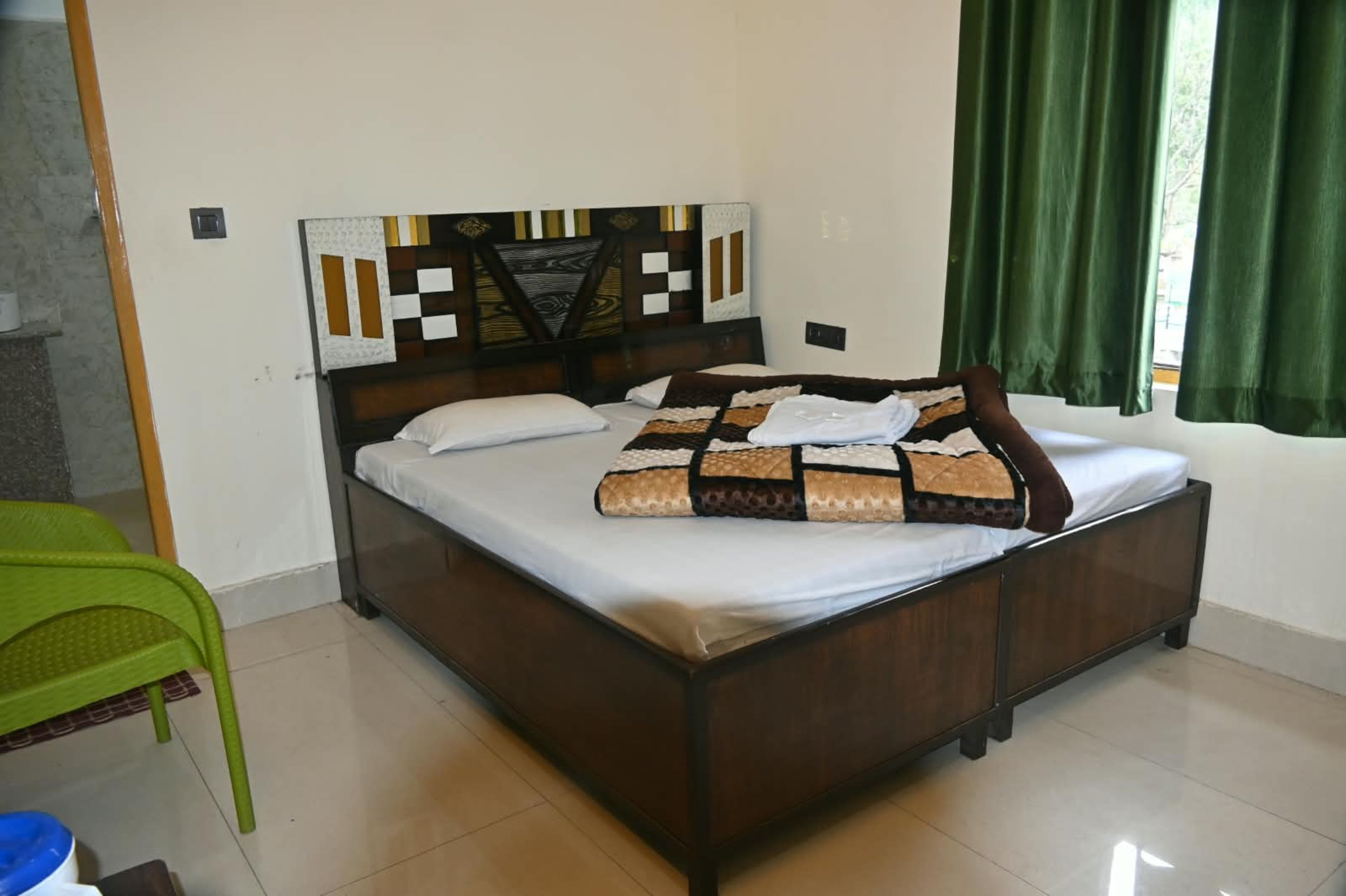 A single bed with a patterned headboard, a green chair, and a window with green curtains.