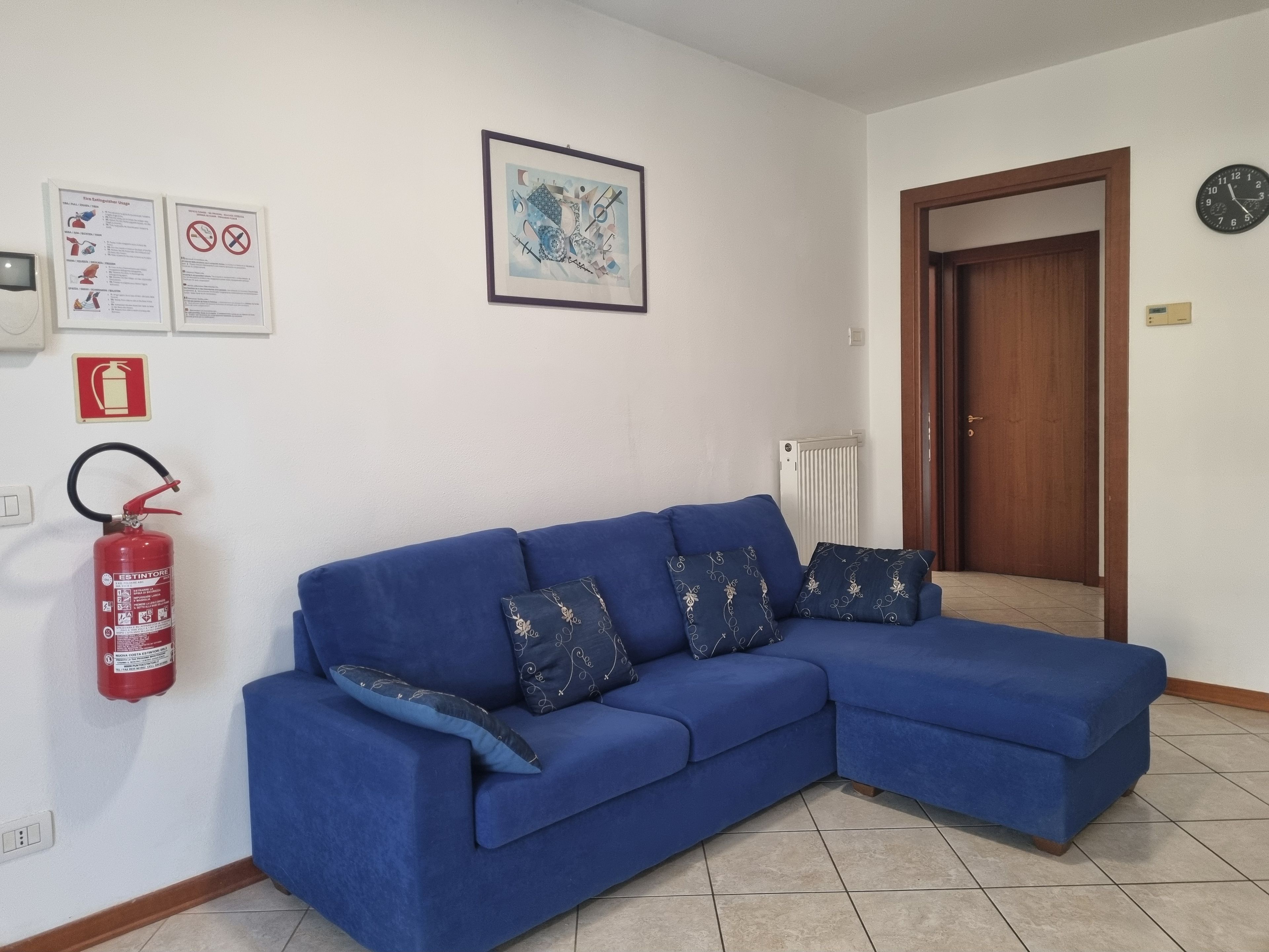Un sofá azul con cojines estampados en una habitación que incluye un extintor, una obra de arte abstracta enmarcada y una puerta.
