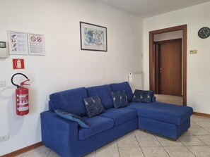 Un sofá azul con cojines estampados en una habitación que incluye un extintor, una obra de arte abstracta enmarcada y una puerta.