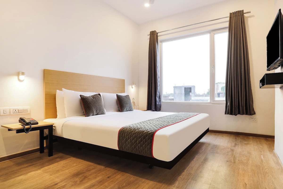 A hotel room with a large bed, a bedside table with a telephone, a window with curtains, and a flat-screen TV mounted on the wall.