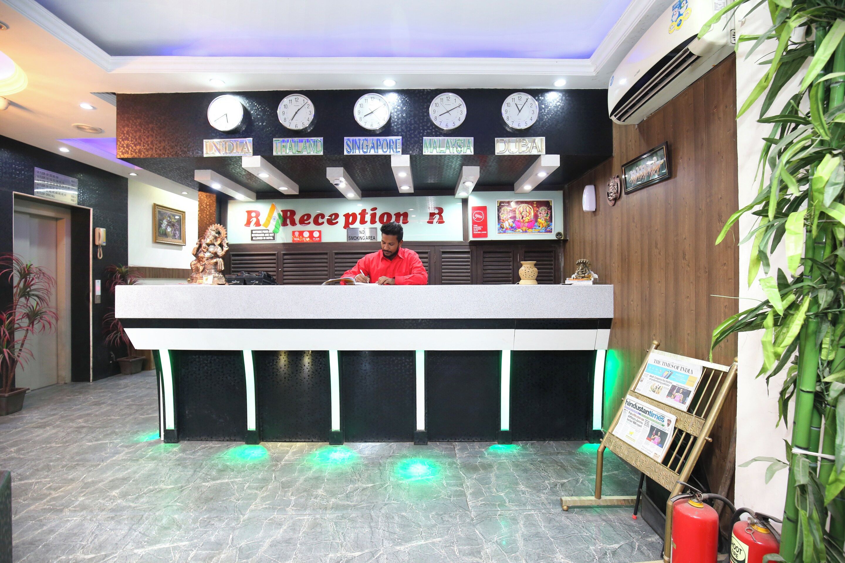 Una recepción de hotel con relojes que muestran diferentes husos horarios, un cartel de recepción y un extintor a la derecha.