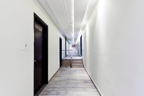 Ein langer, schmaler Korridor mit einer Reihe von Türen an einer Seite, einer Treppe am Ende und einer Leuchtkette an der Decke.