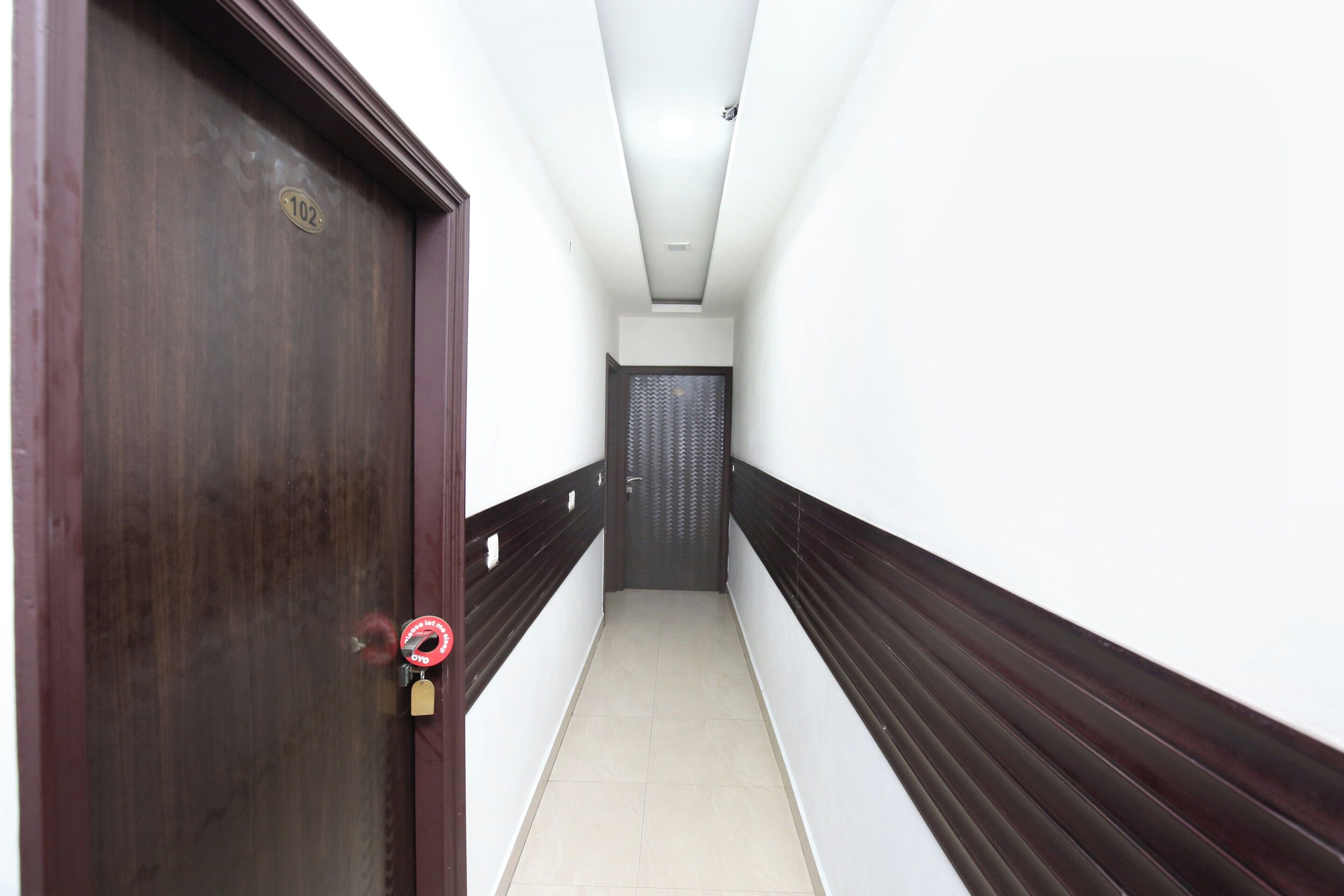 Um corredor com uma porta de madeira à esquerda, uma porta preta no final e paredes brancas com lambris nas laterais.