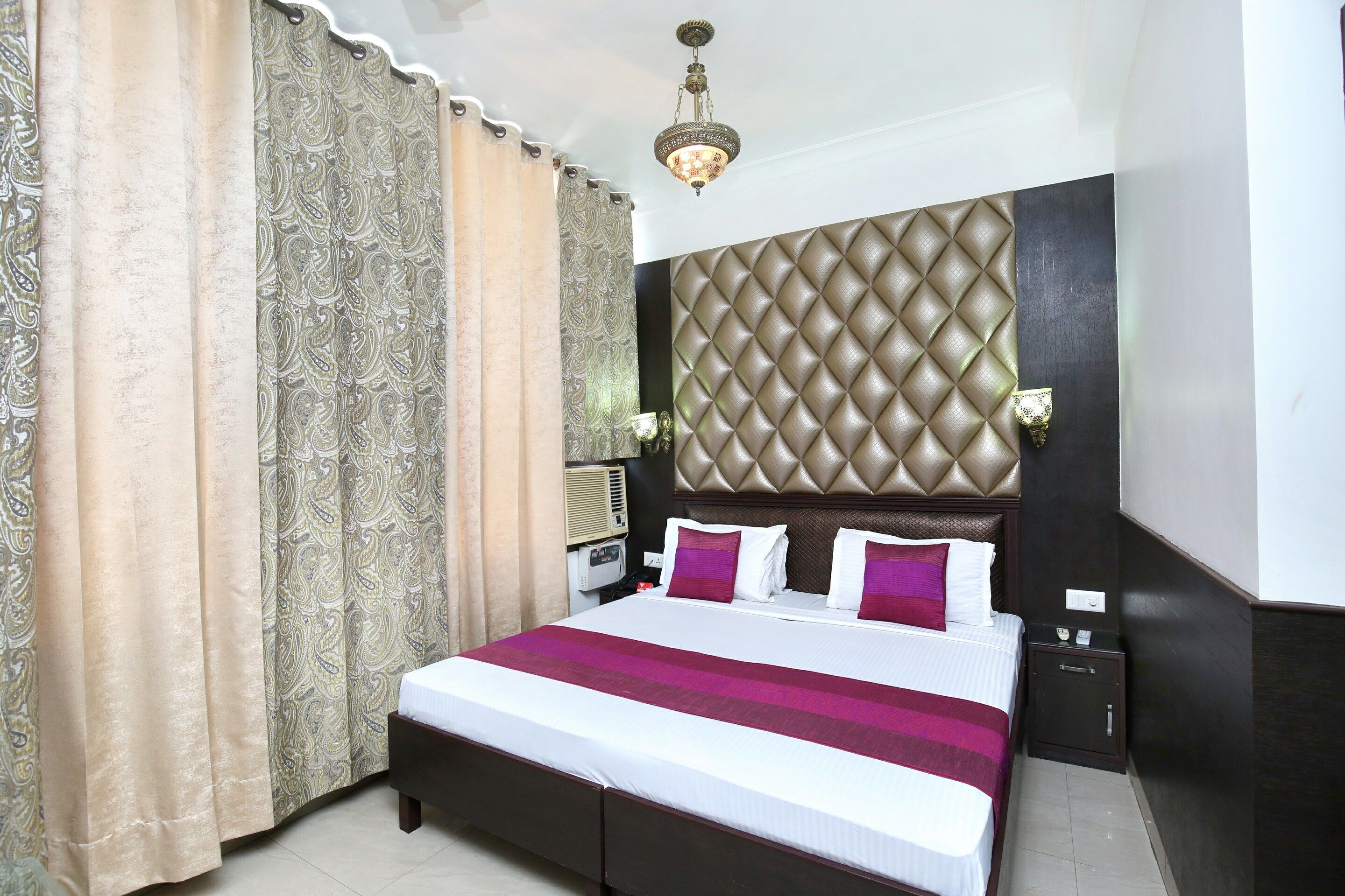 A hotel room with a large bed, a headboard, curtains, a nightstand, and a wall-mounted light fixture.