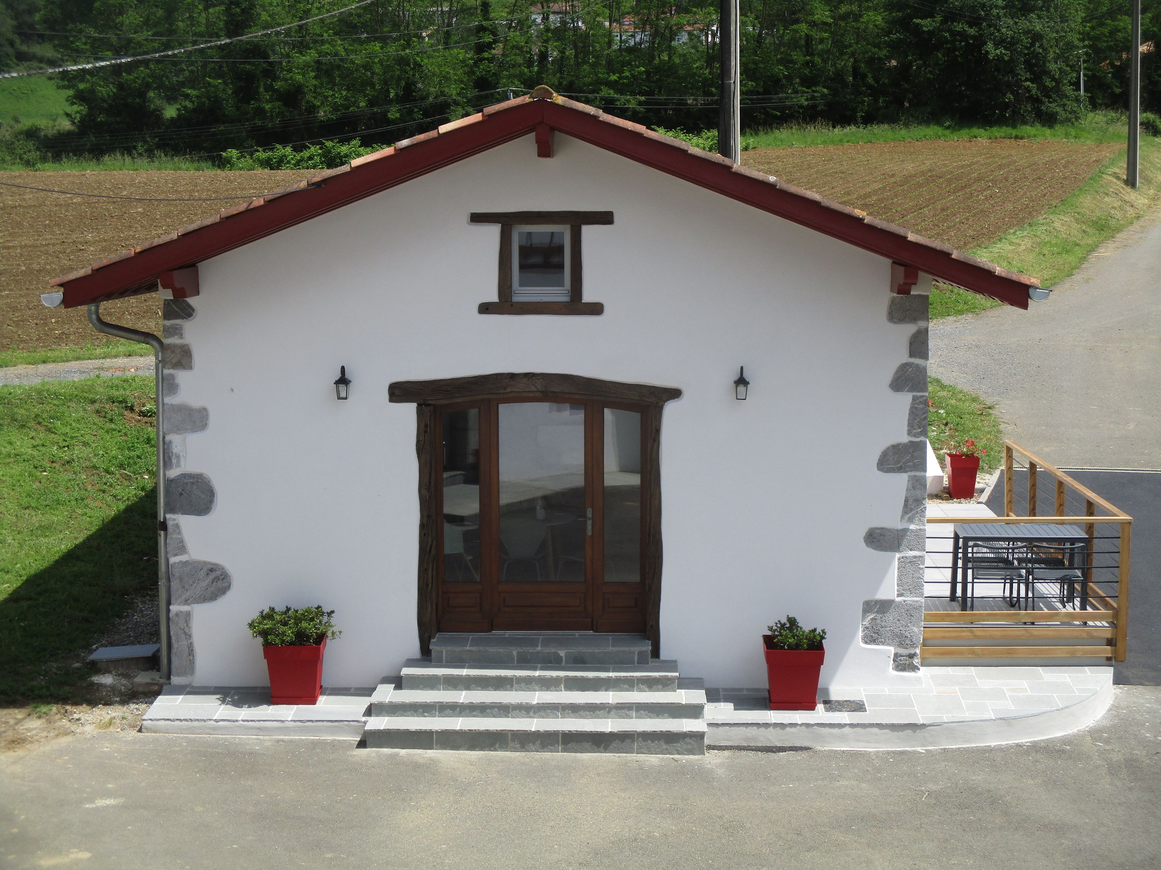A small, single-story building with a red roof, a wooden door, and two potted plants on the ground floor.