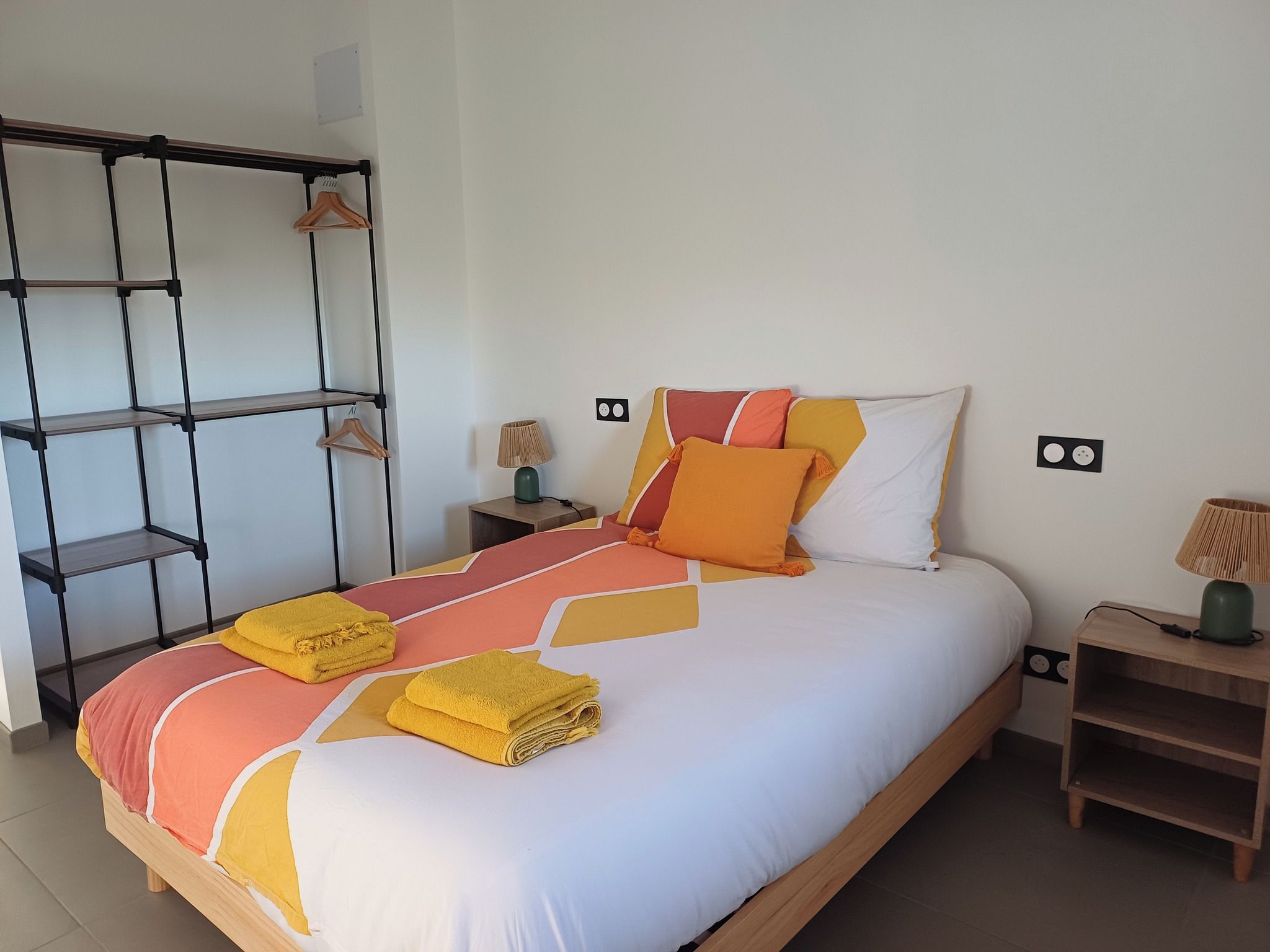 Un letto rifatto con cura, coperta a motivi geometrici, due comodini con lampade e una mensola a muro con appendiabiti.