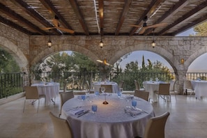 Un área de comedor al aire libre con arcos de piedra, mesas redondas y sillas, bajo un techo de madera con luces colgantes.