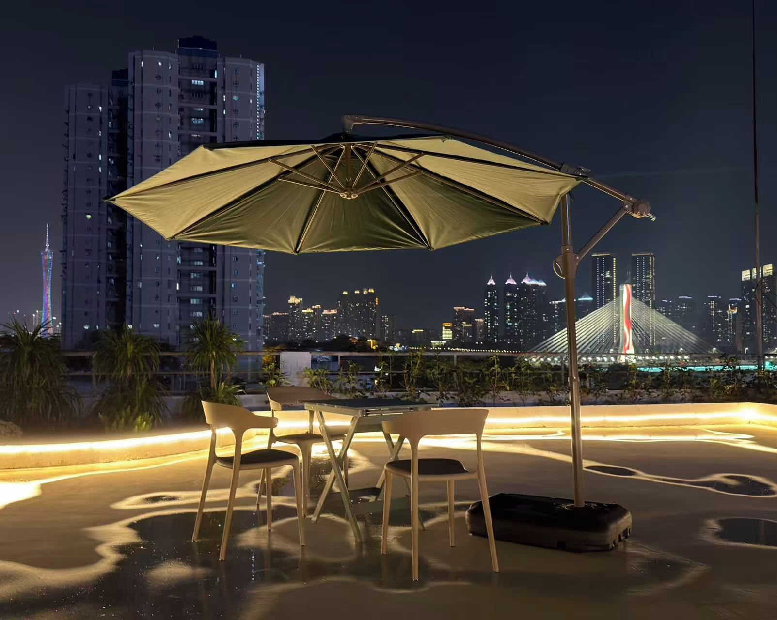 An outdoor seating area with a table and chairs under a large umbrella, overlooking a city skyline with illuminated buildings and a bridge.