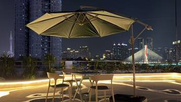 An outdoor seating area with a table and chairs under a large umbrella, overlooking a city skyline with illuminated buildings and a bridge.