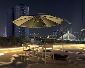An outdoor seating area with a table and chairs under a large umbrella, overlooking a city skyline with illuminated buildings and a bridge. - Sunrise Voyage (Guangzhou)
